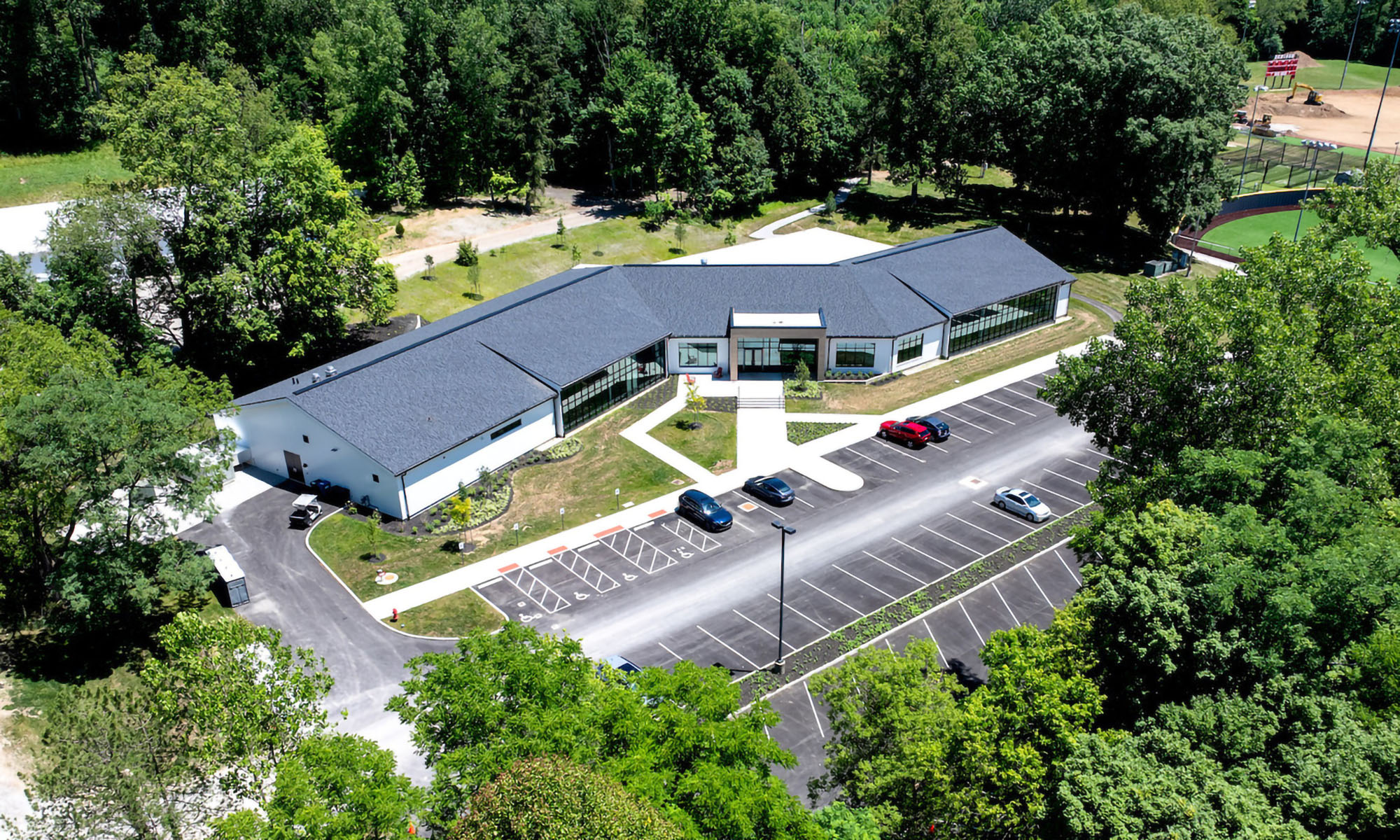 Aerial view of a single-story building with a dark roof, surrounded by trees, a parking lot with several cars, and a walkway leading to the entrance.