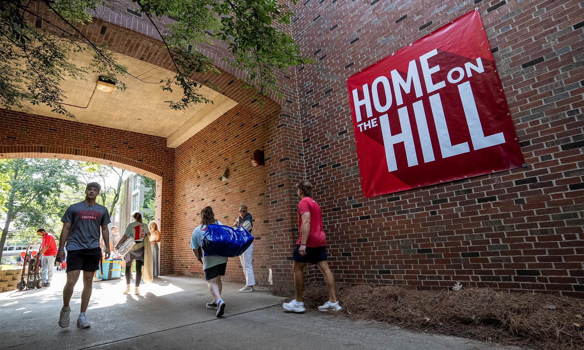 Students and families carrying bags and items through the archway of a large brick residence hall displaying a giant red and white banner that says "Home on The Hill."