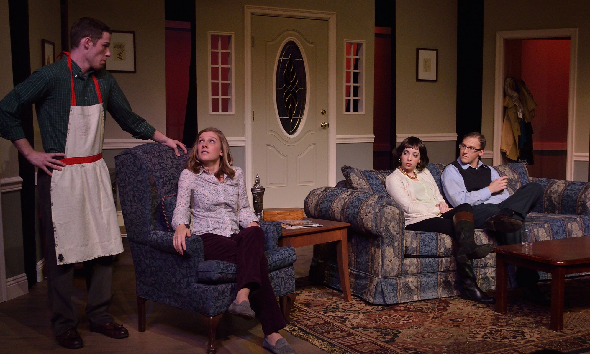 Ken, wearing a feminine apron, leans on the armchair Hannah is seating in. Hannah looks up to him as Beth and John sit on the couch and look at Hannah.Ken, wearing a feminine apron, leans on the armchair Hannah is seating in. Hannah looks up to him as Beth and John sit on the couch and look at Hannah.