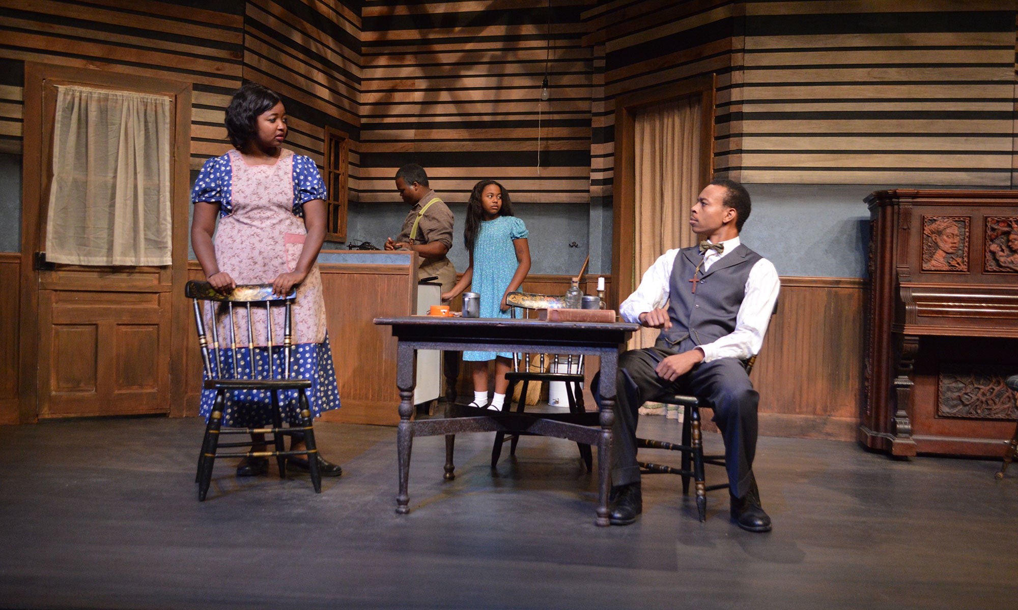 Avery sits at the table, looking at Berniece. Behind them, Doaker cooks in the kitchen as Maretha looks toward the family piano.
