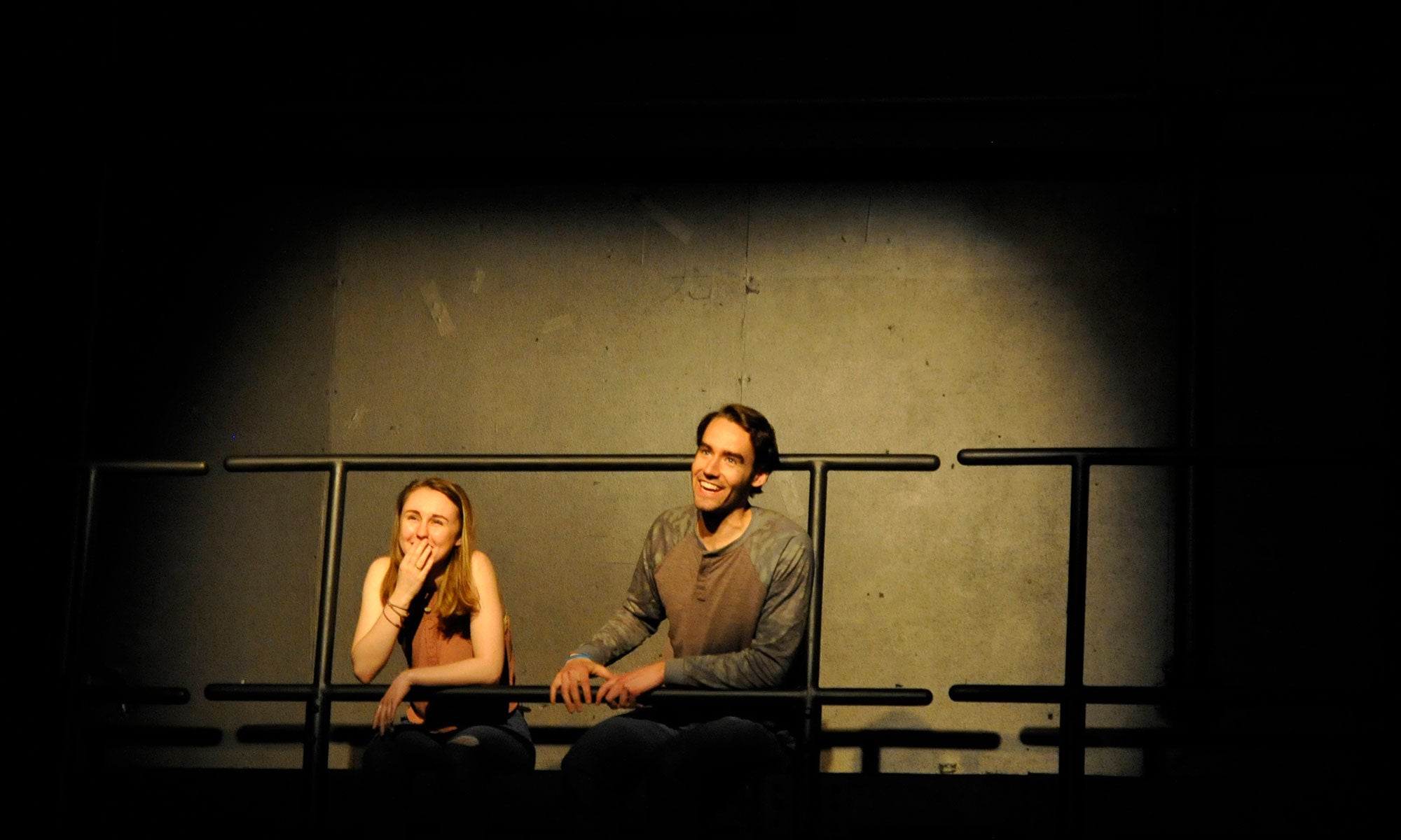 Two actors laugh as they sit off the ledge of the upper level of the Hylbert Family Studio Theatre, performing the short play Baby Steps.