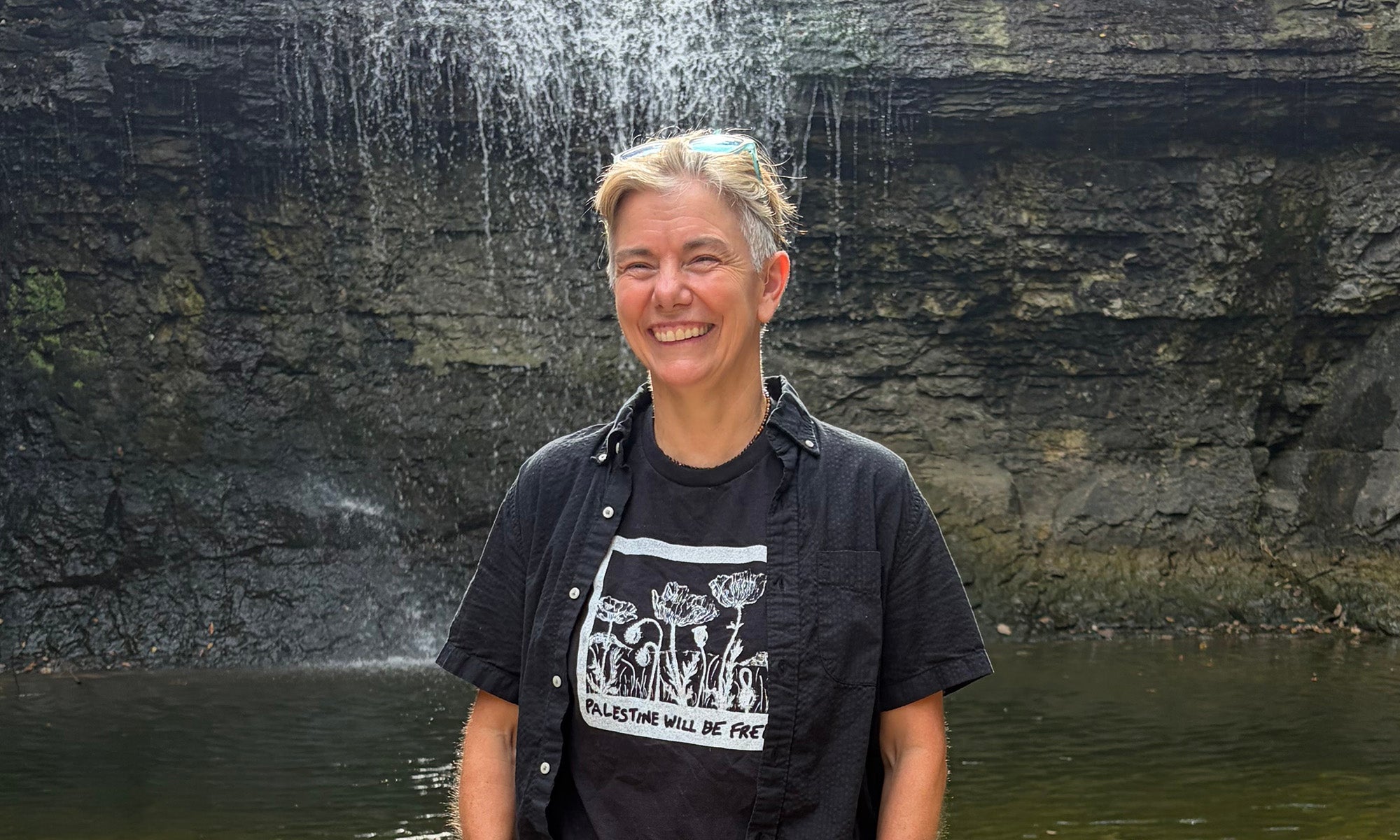 Person standing and smiling in front of a small waterfall, wearing a black shirt and sunglasses on their head.