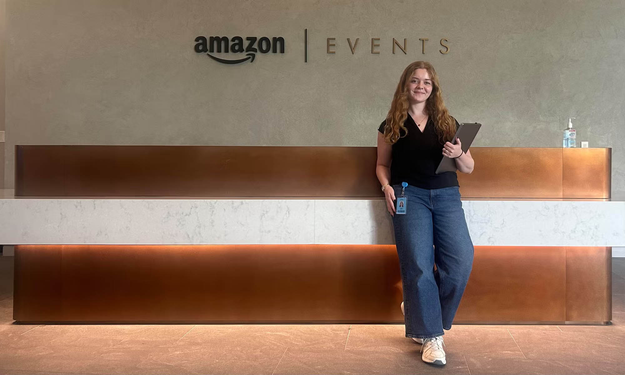 A woman with long reddish blonde hair in professional clothing stands in front of a large modern reception desk holding a laptop. Behind her, the Amazon logo next to the word "Events" hands on a textured taupe-colored wall.