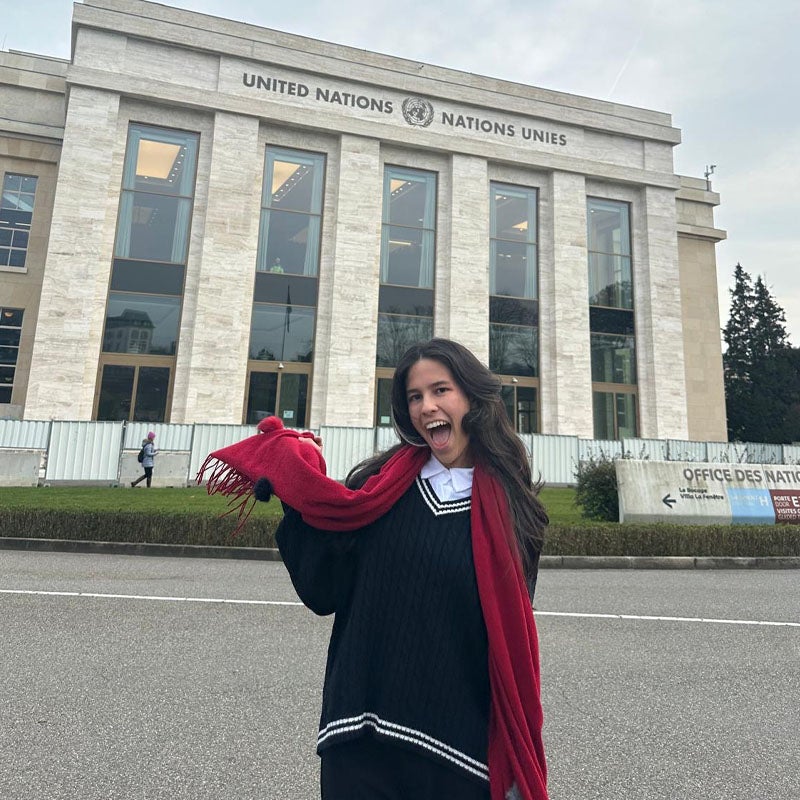 Anna Laura Ragazzoni Xavier Batista  A woman in a black sweater and long red scarf smiles excitedly for the camera while standing in front a United Nations building.
