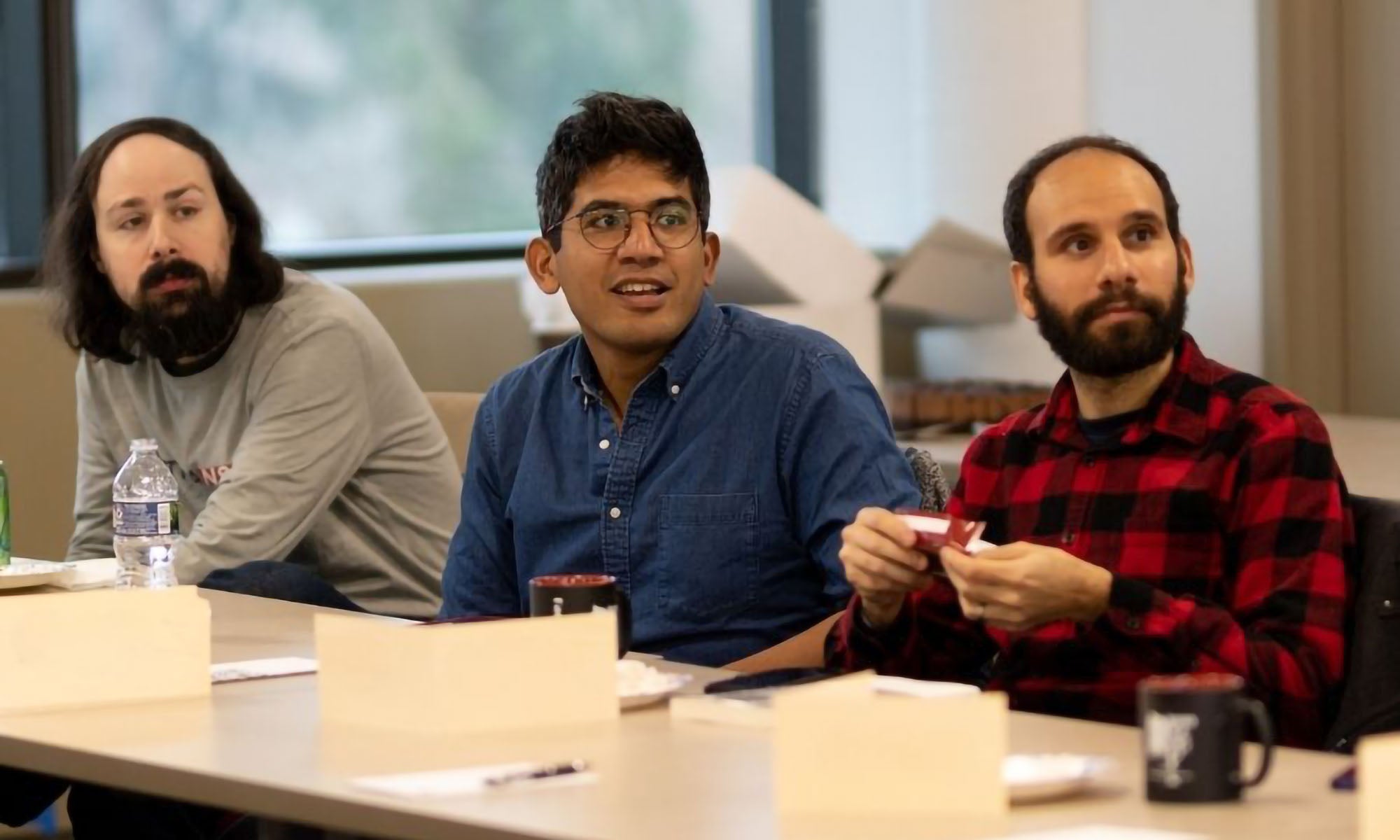 Three men sit at a table in a classroom style conference room listening to a presentation.