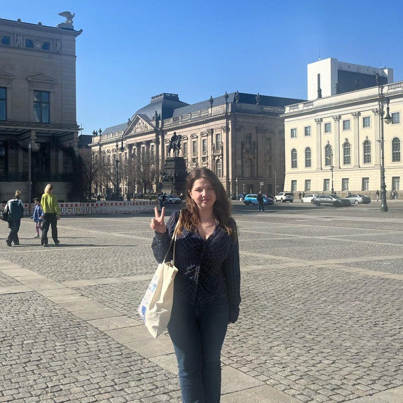 Avery Maurer A woman in blue plaid shirt and jeans, carrying a tote on her shoulder, stands in a city plaza surrounded by ornate stone buildings, holding her right hand in a traditional "peace sign" symbol.
