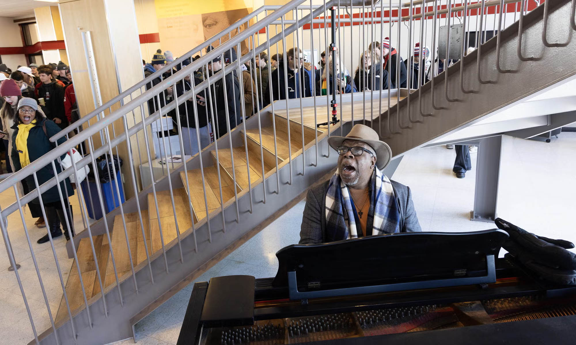 A man in a hat wearing a suit coat and scarf plays a piano in front of a modern staircase as a  large group of people listen.