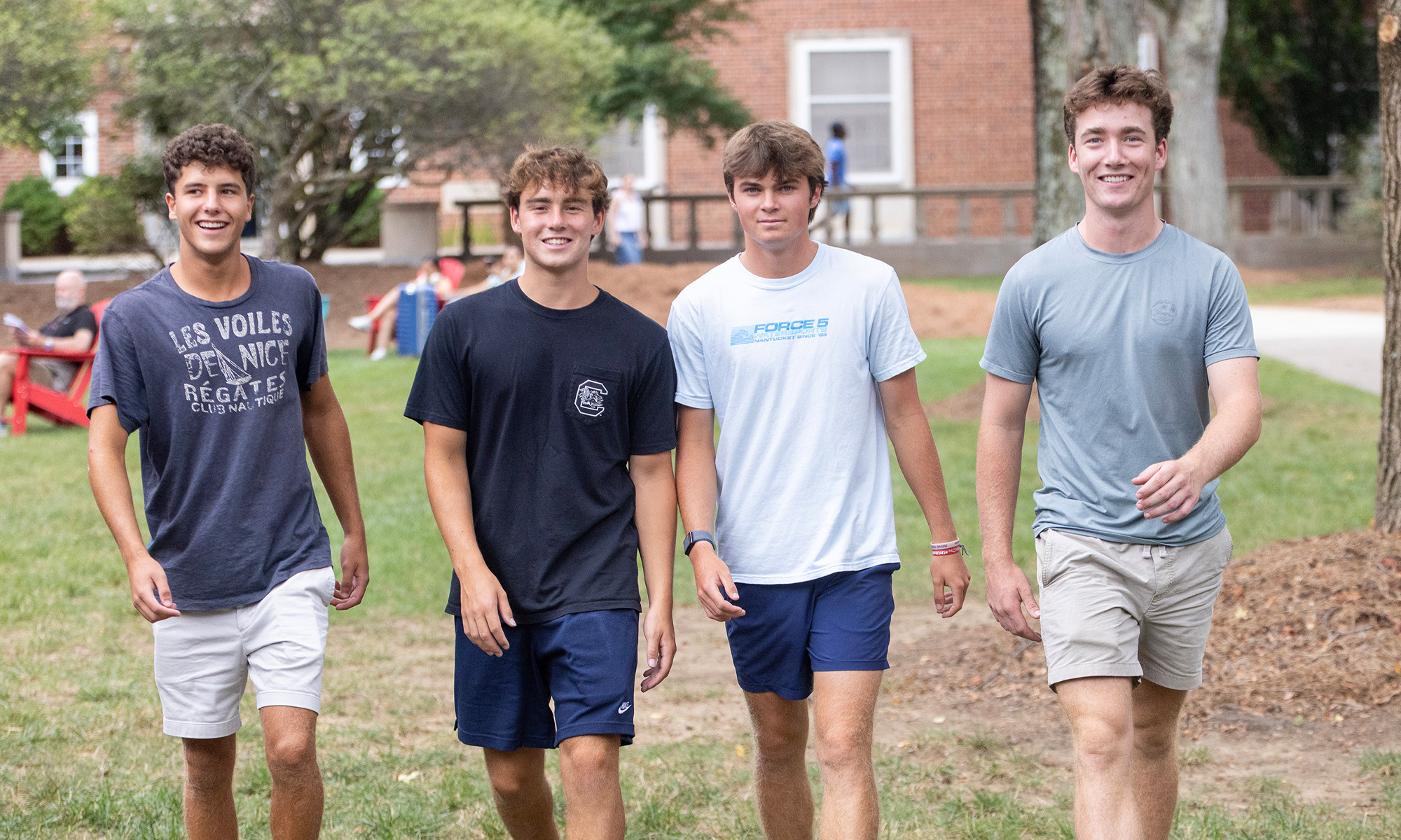 Four college students walking toward the camera. In the background, other students and families can be seen relaxing on the quad.