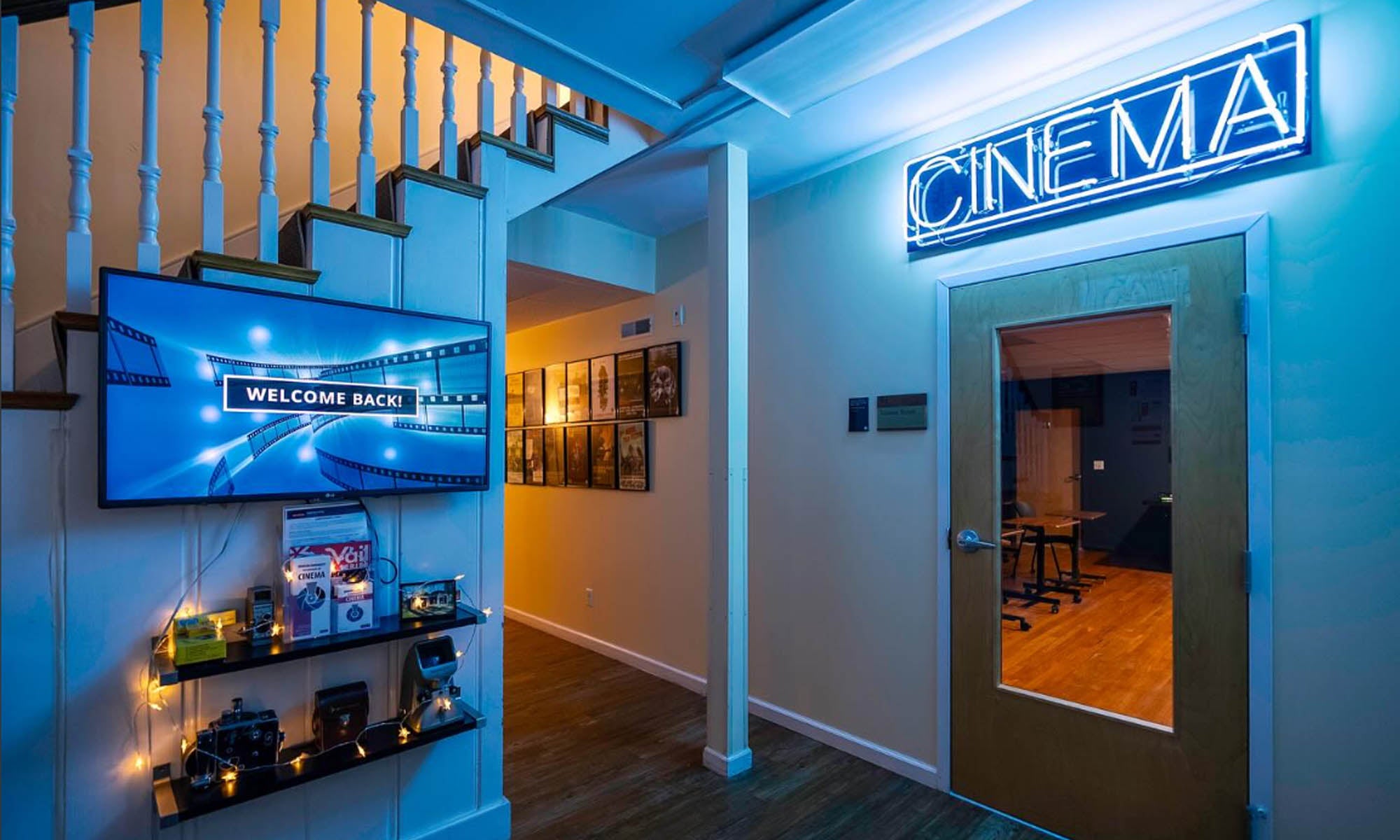 A house interior with a neon sign that reads "Cinema" in capital letters above a wooden door with a glass window. A display of cinema-related equipment and flat-screen television mounted on a nearby staircase is on the left next to a hallway entrance.