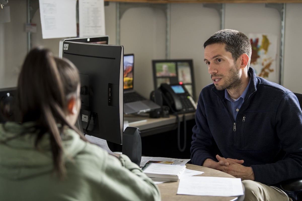 Two people sit across a desk in an office, engaged in conversation