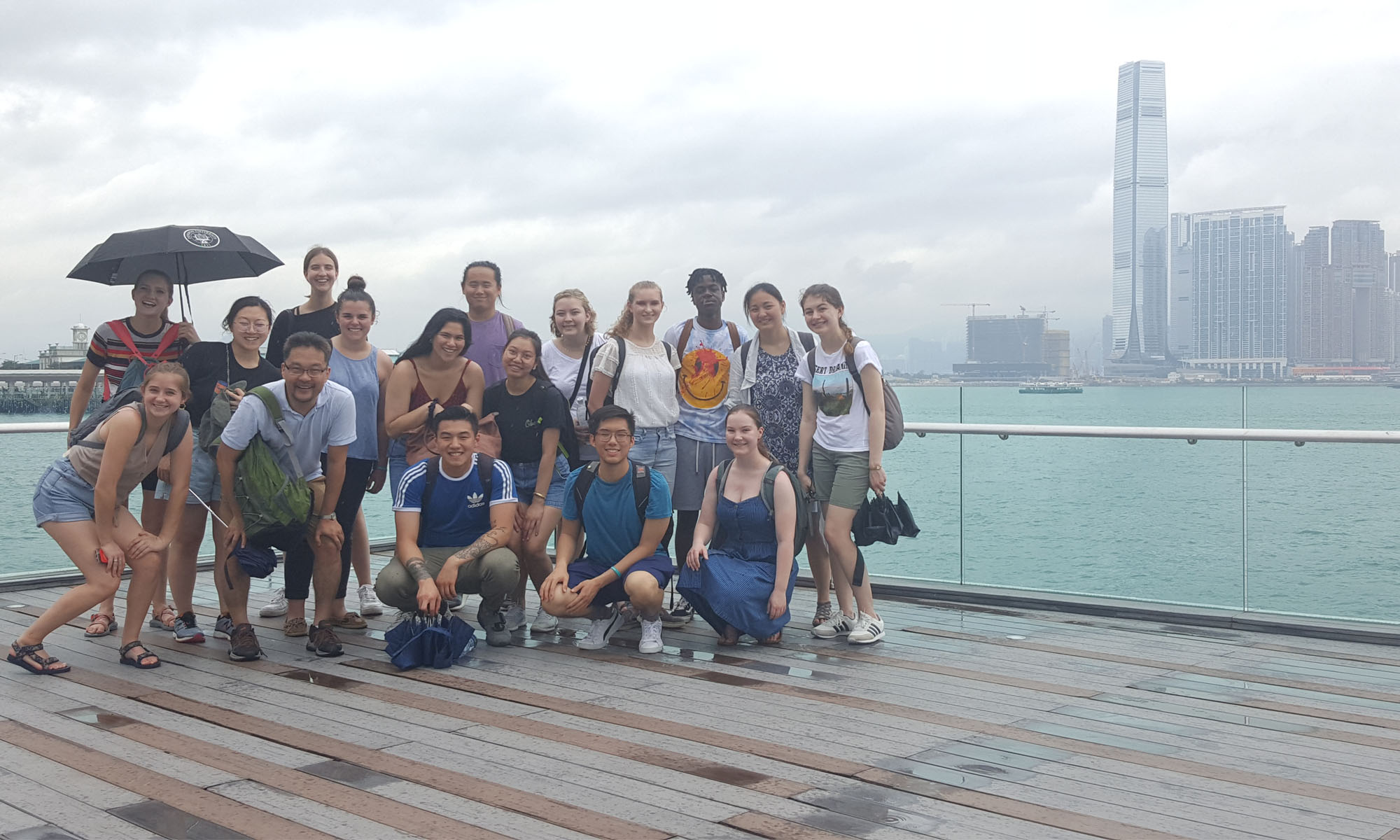 A large group of students posing for a photo on a sea port deck. There is a glass wall with a metal railing behind them that overlooks a bay of water with skyscrapers in the distance.