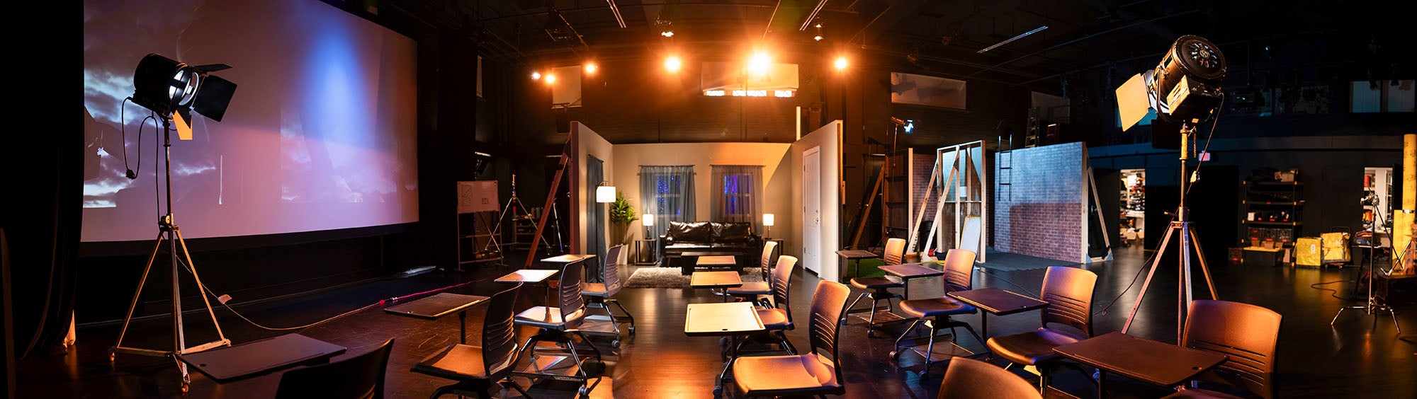 A panoramic photo of class room desks set up in a film studio facing a large projection screen, surrounded by lighting equipment, set pieces, and production sets.