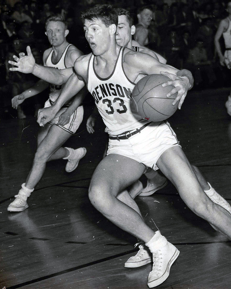 Black and white photo of Don DeJong '54 playing basketball at Denison; he wears a white uniform with dark trim, sporting the number 33, and is mid-dribble, running down the court.