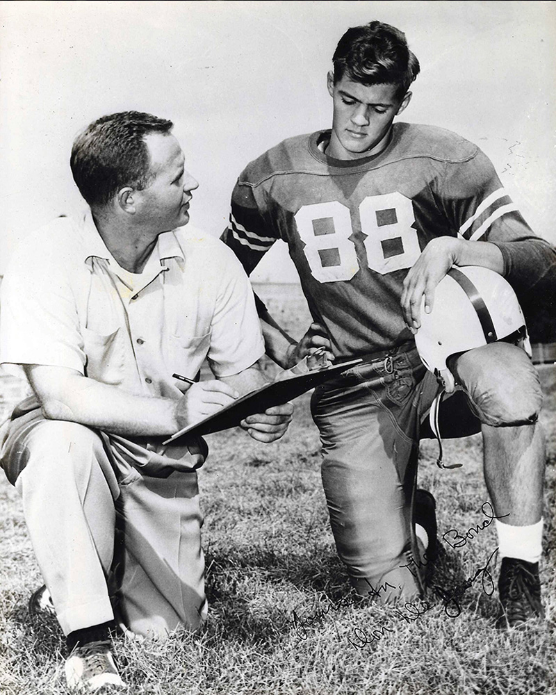 Black and white photo of Don DeJong in a Denison football uniform reviewing a play with a member of the coaching staff.