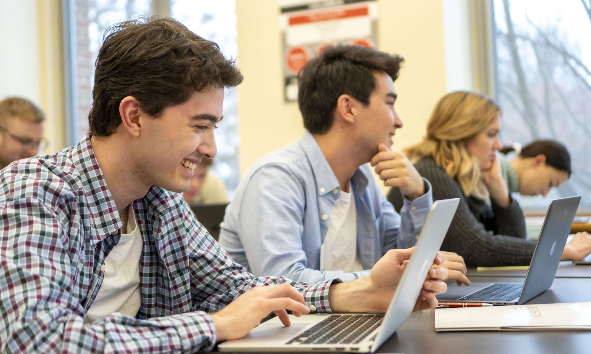 A classroom of students sitting at tables with their laptops, taking notes and listening to their professor.