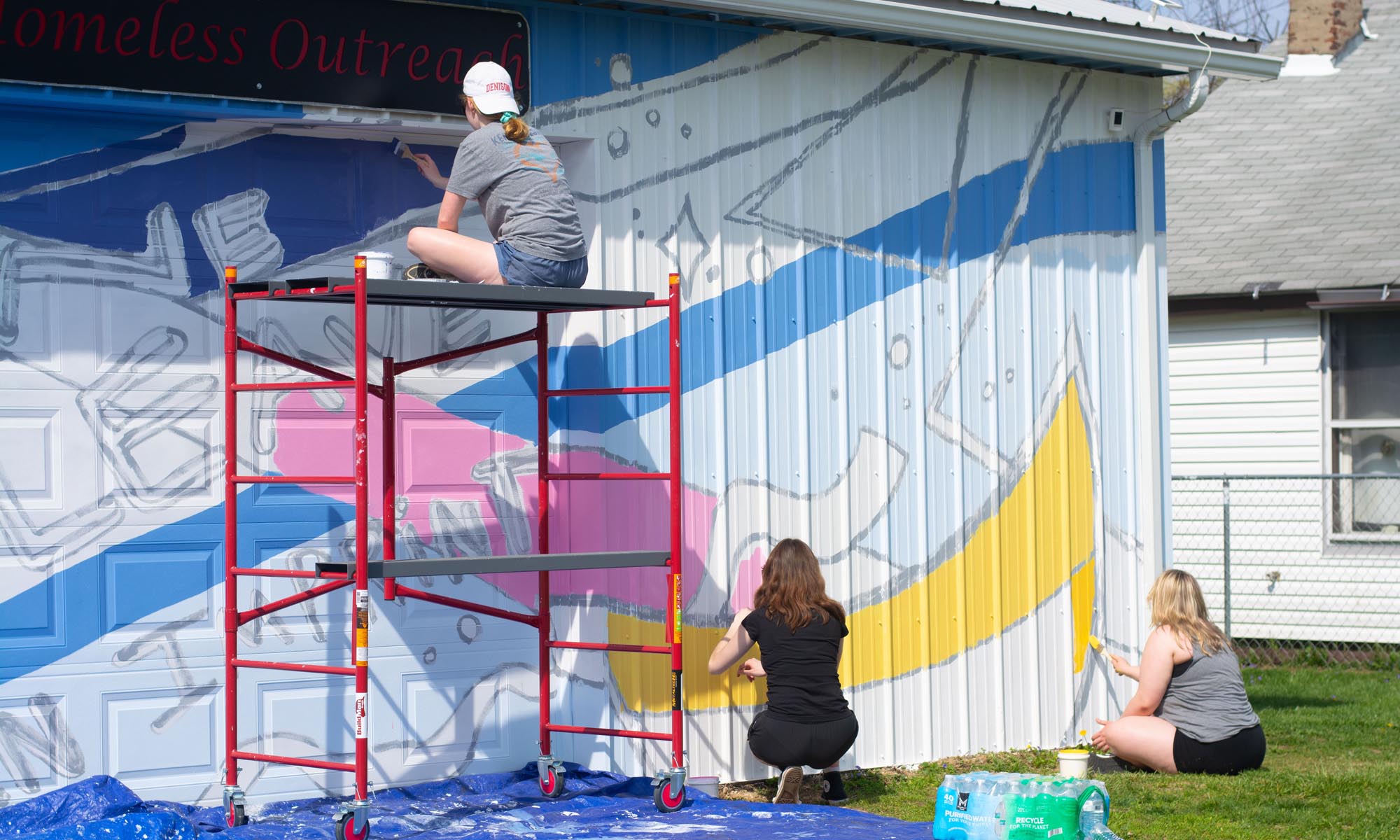 Student artists and volunteers painting a mural on the side of a building.