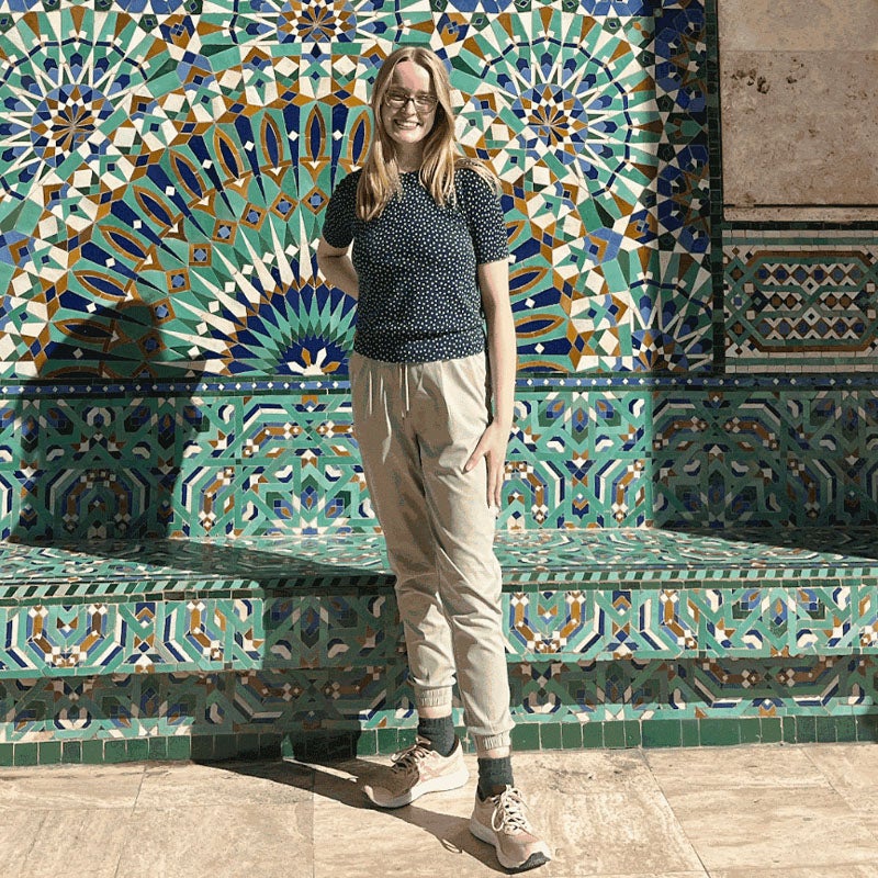 Eliza Scoggin  A woman wearing a navy-blue shirt with small white polka dots and beige pants stands in front of a mosaic tiled wall.
