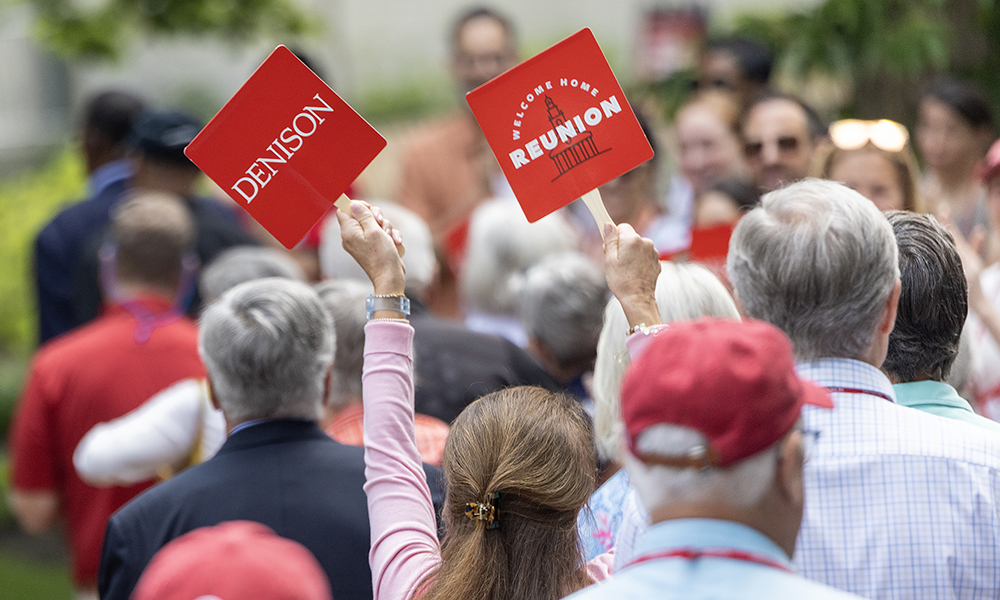A photo of dozens of alumni filling the frame, walking together; a woman holds up a two small signs that say "Denison" and "Reunion 2025."
