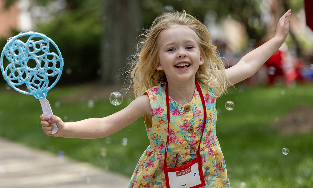 A young girl — a future Denisonian? — holds a large blue bubble wand and runs through bubbles on the quad.