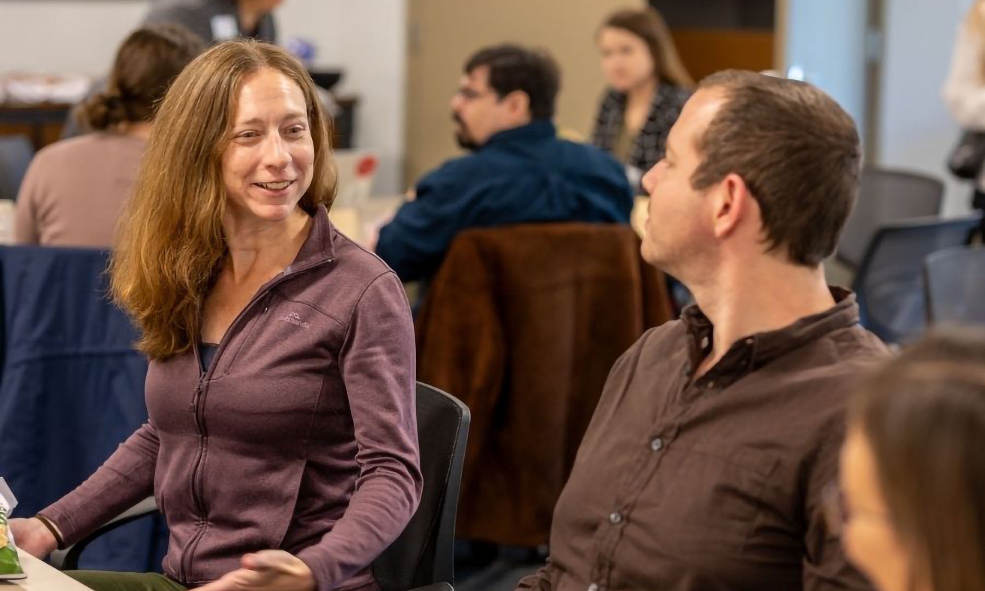 A woman with long light brown hair talks to a man with brown hair, both in business casual wear, in a conference room.