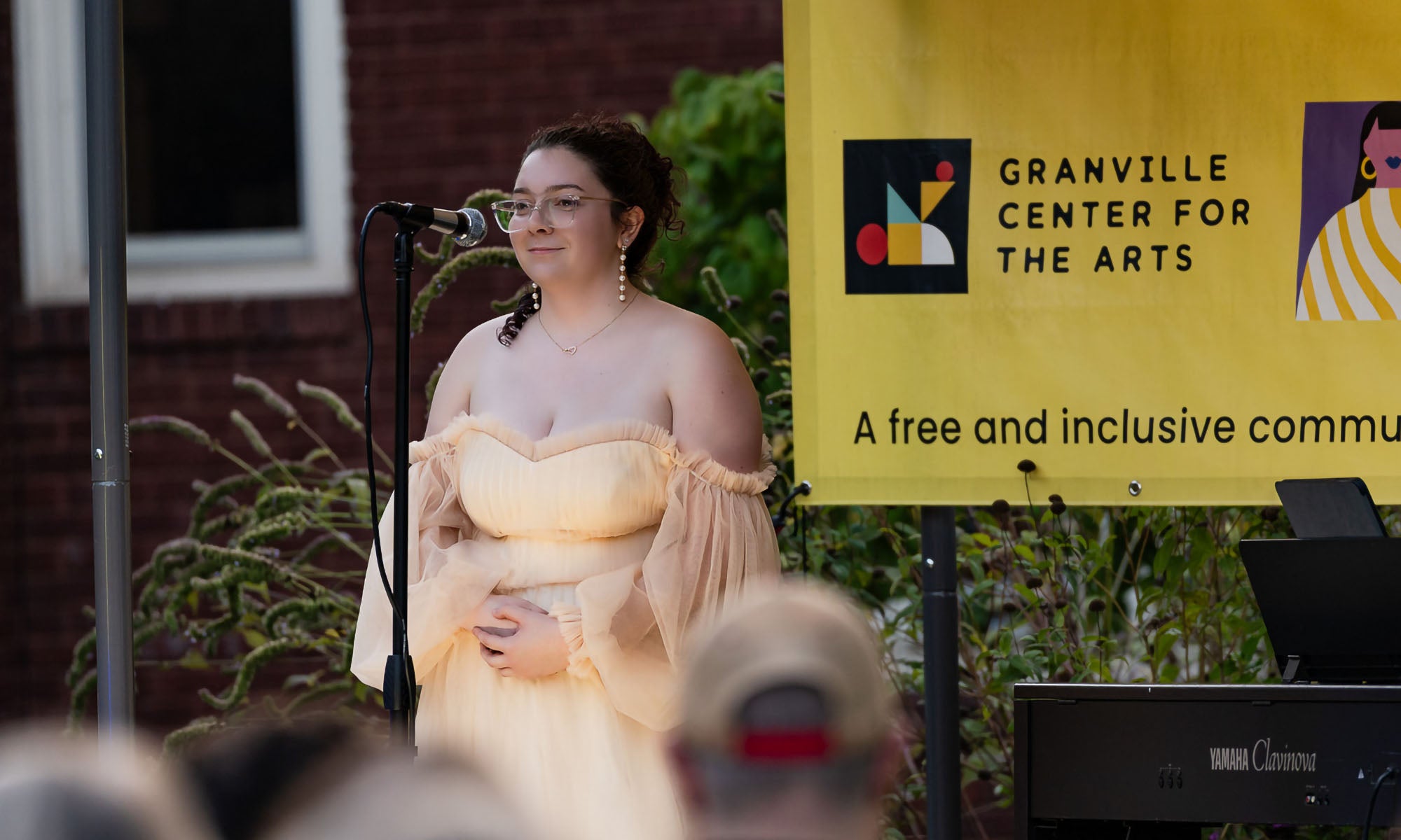 Gabby Belmonte ’27 at the Granville Center for the Arts. A woman in a light, off-the-shoulder dress stands at a microphone on an outdoor stage beside a yellow Granville Center for the Arts sign.