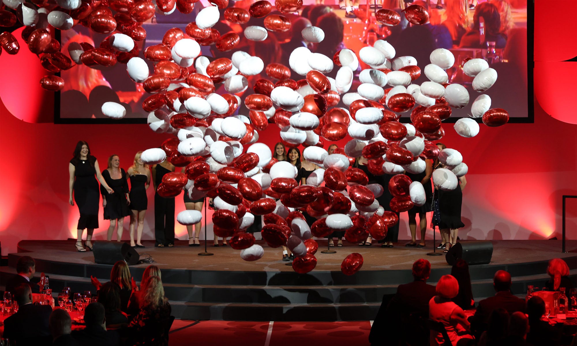 Hundreds of red and white balloons fall from the ceiling above a stage illuminated with red lights during an event in a large event space.