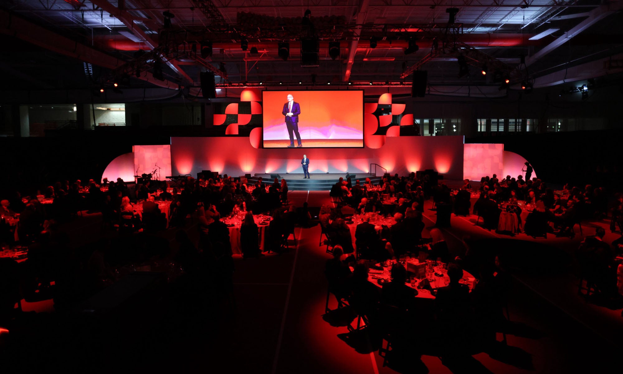 A person stands on a stage giving a presentation to an audience seated at round tables, with red lighting illuminating the event space.