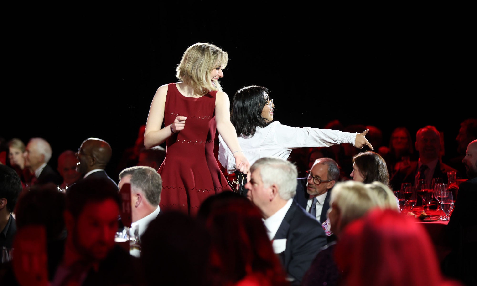 A blonde woman in a red dress and a woman with dark hair wearing white perform a song surrounded by people sitting at tables during an event.