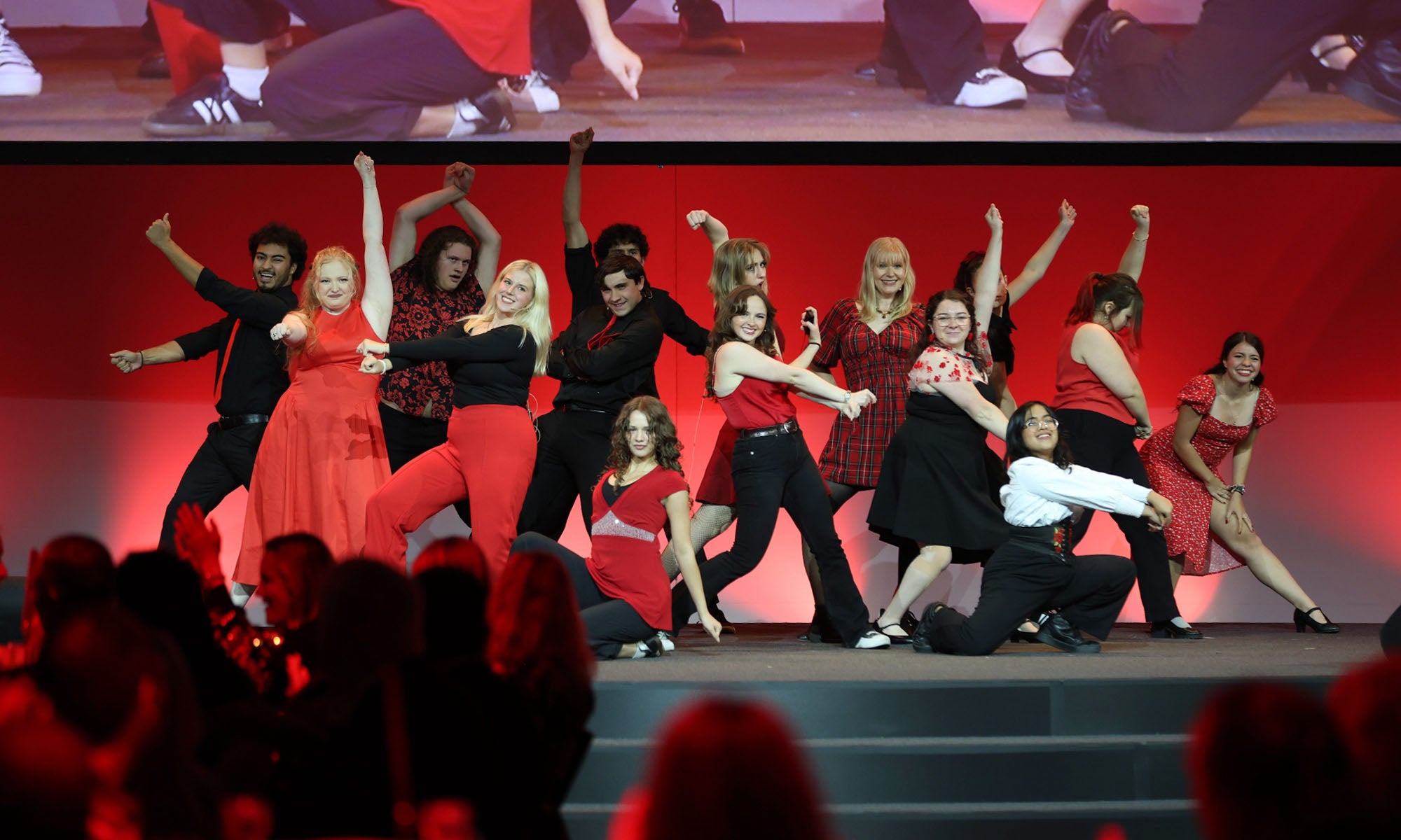 A large group of performers wearing combinations of red, black, and white, strike a pose during a performance on a stage at a large event.