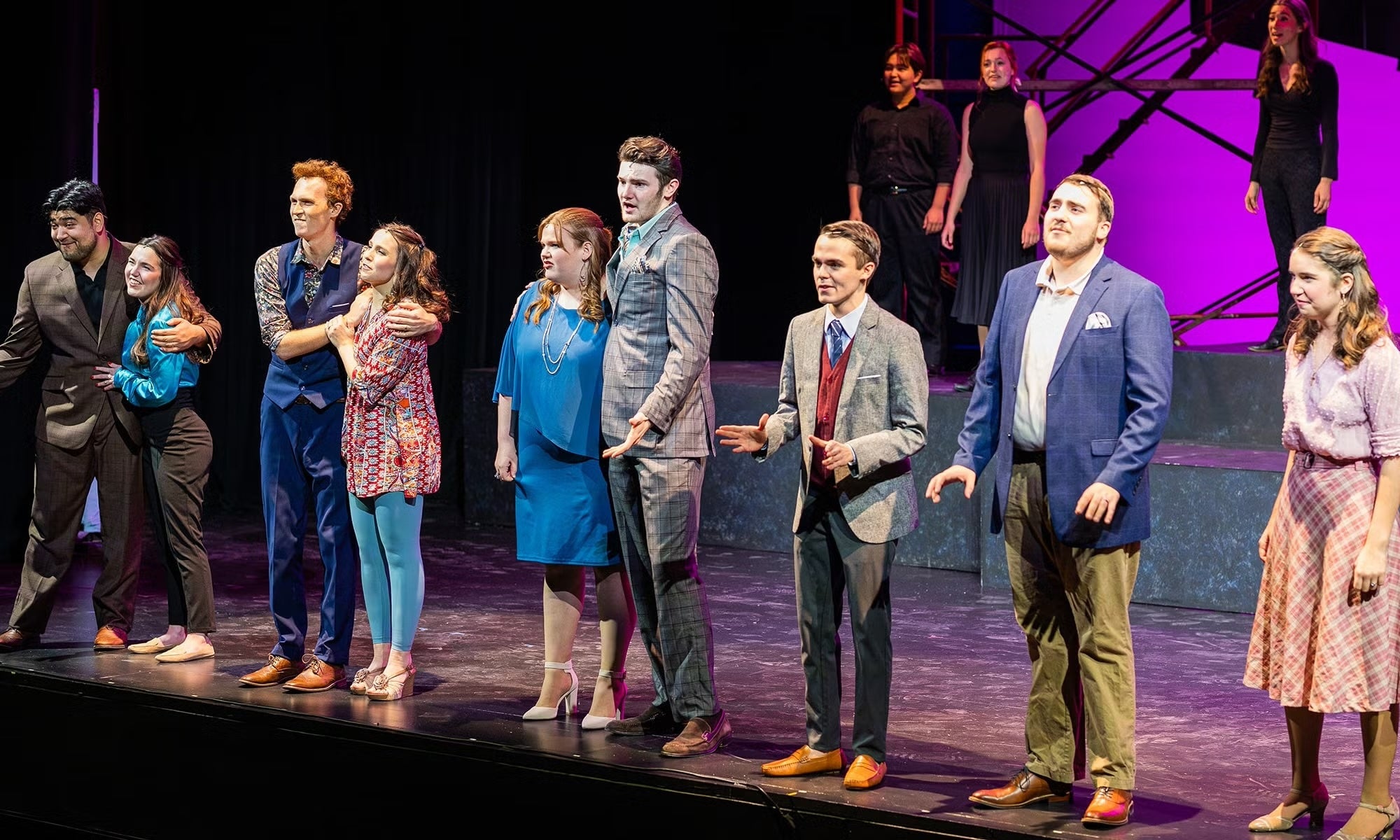 A group of actors in formal and semi-formalwear stand on stage during a musical theater performance