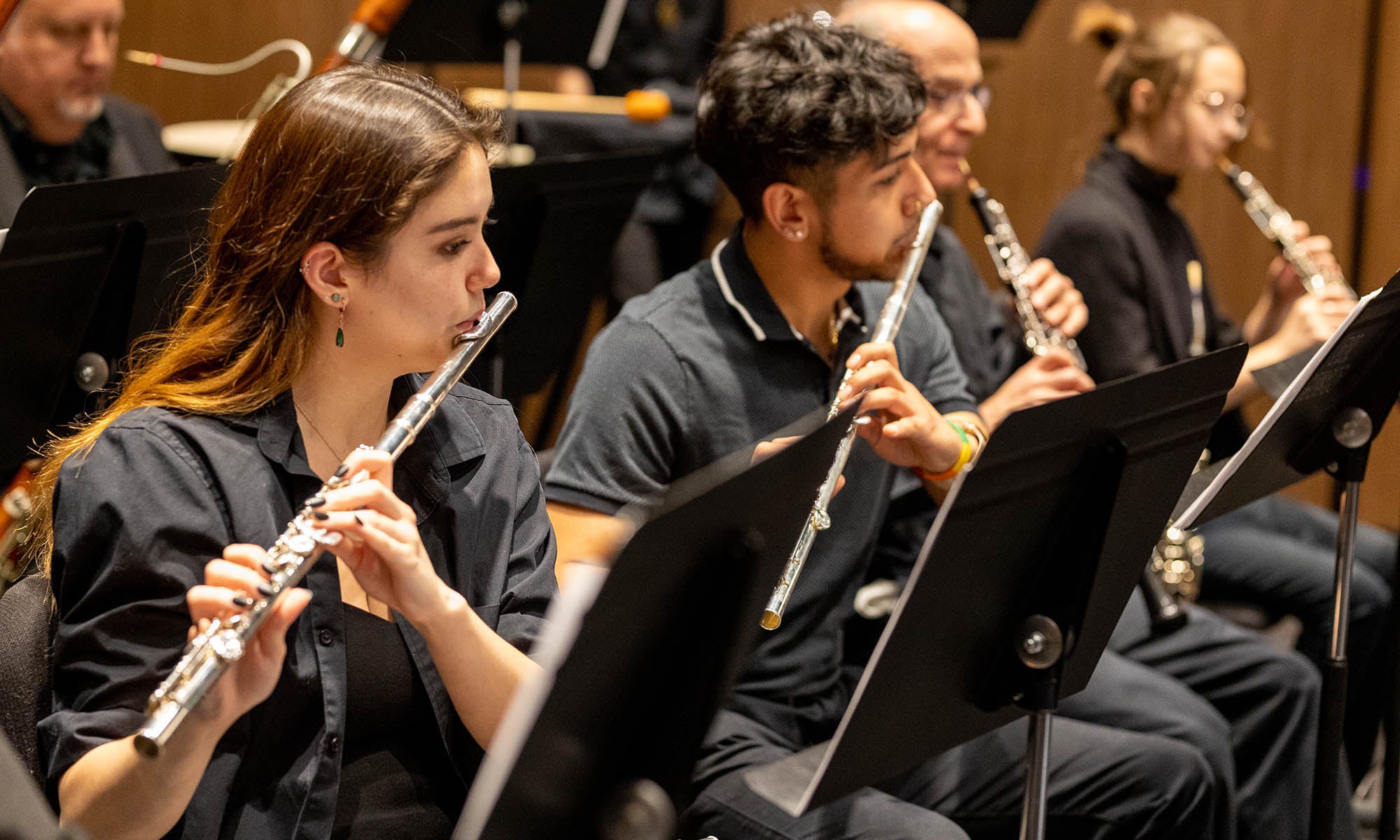 Students sitting in front of music stands playing flutes, oboes, and other instruments in concert on a stage.