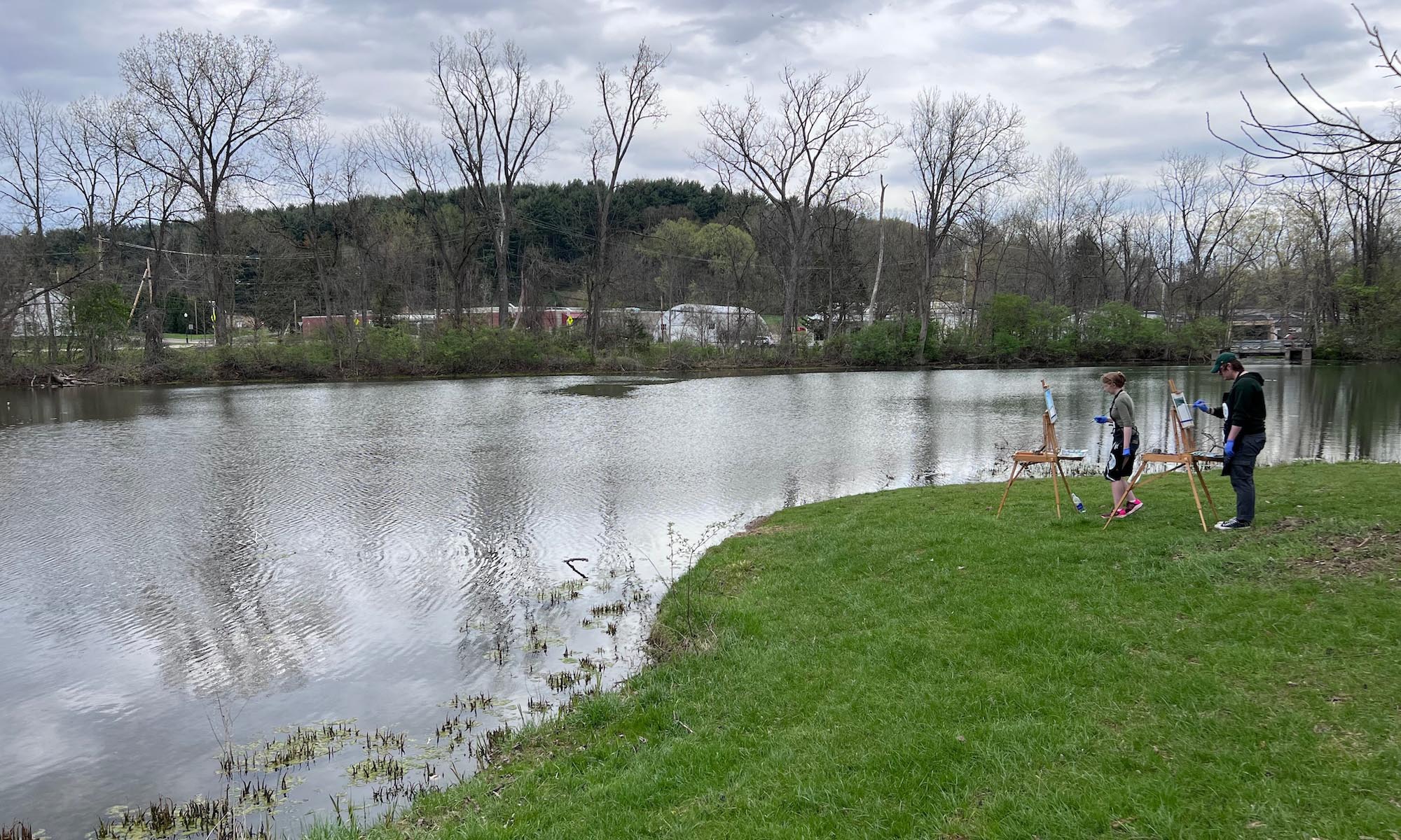 A view of Ebaugh Pond. Two students with easels are painting by the water's edge.