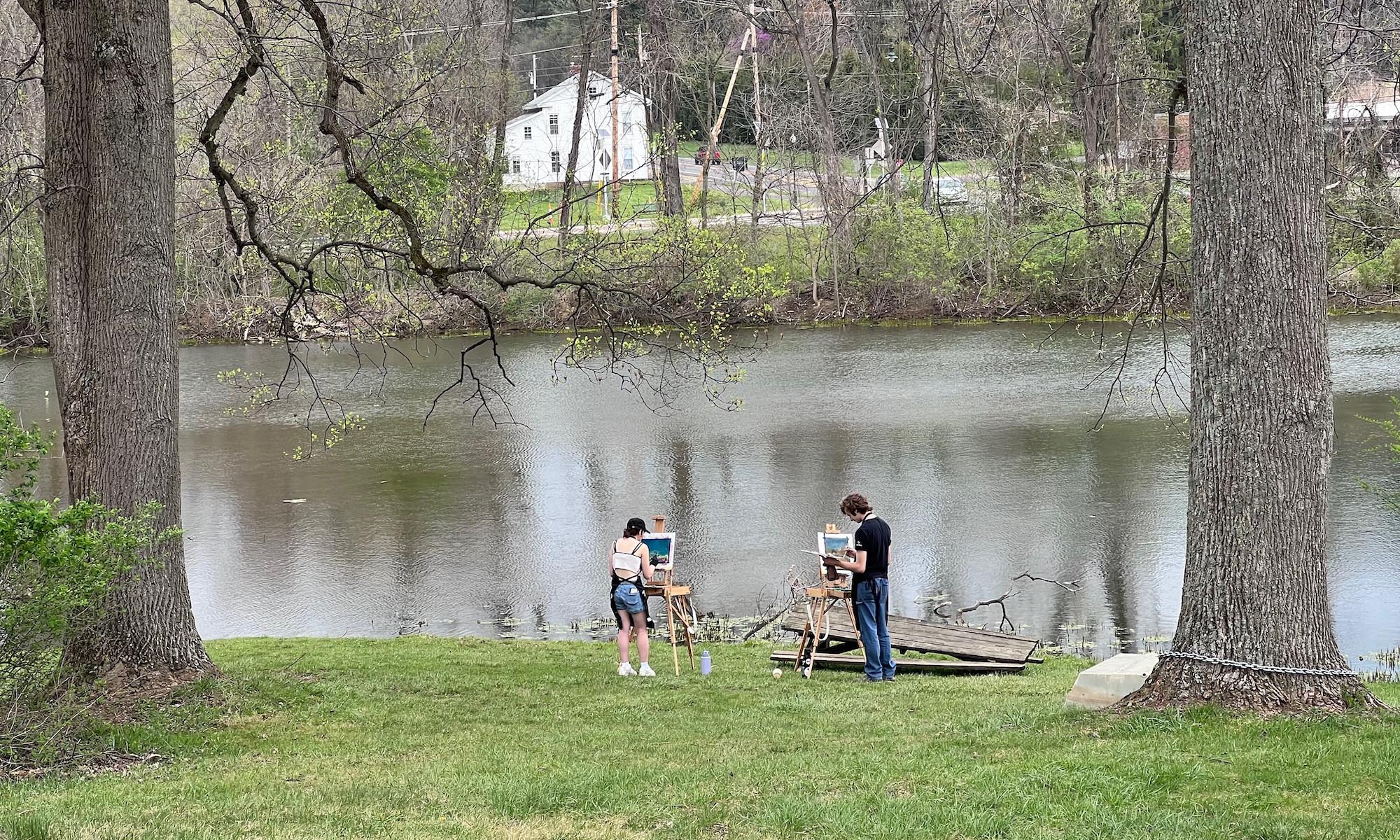 A view of Ebaugh pond from a distance; two trees frame the photo. At the center, several students are painting at easels near the water's edge.