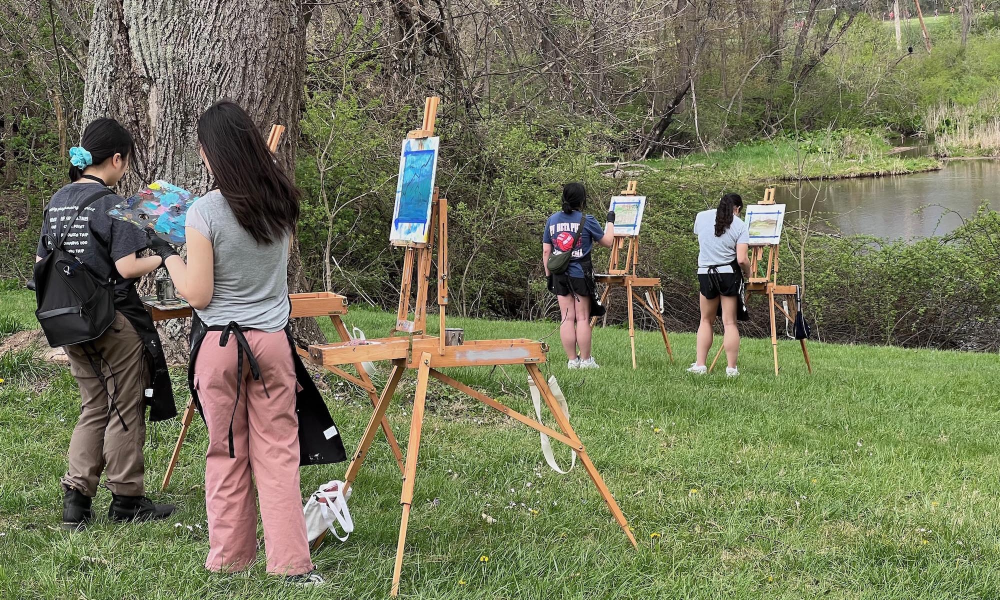 A photo of several Visual Arts students on the banks of Ebaugh pond, all with easels and paint, working on art projects.