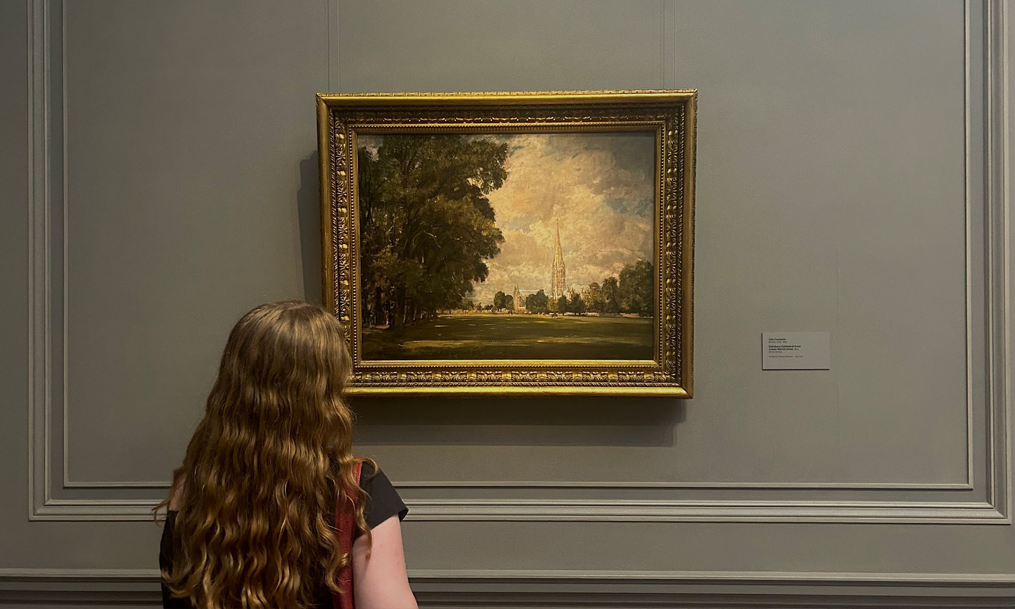 A women with long, wavy reddish hair looks at a painting in a museum.