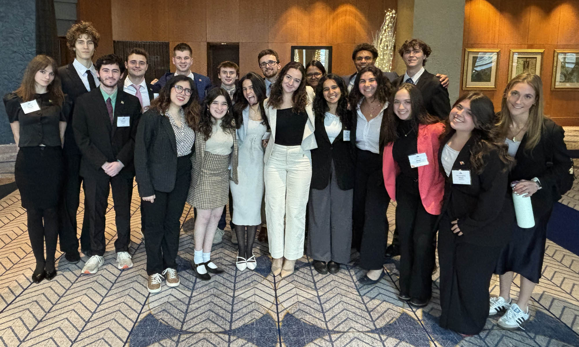 A group of nearly 20 college students in business attire pose for a group photo in the lobby of a conference center that features cream-colored carpet with a dark blue geometric herringbone design, and wood-paneled walls with framed art in the background.