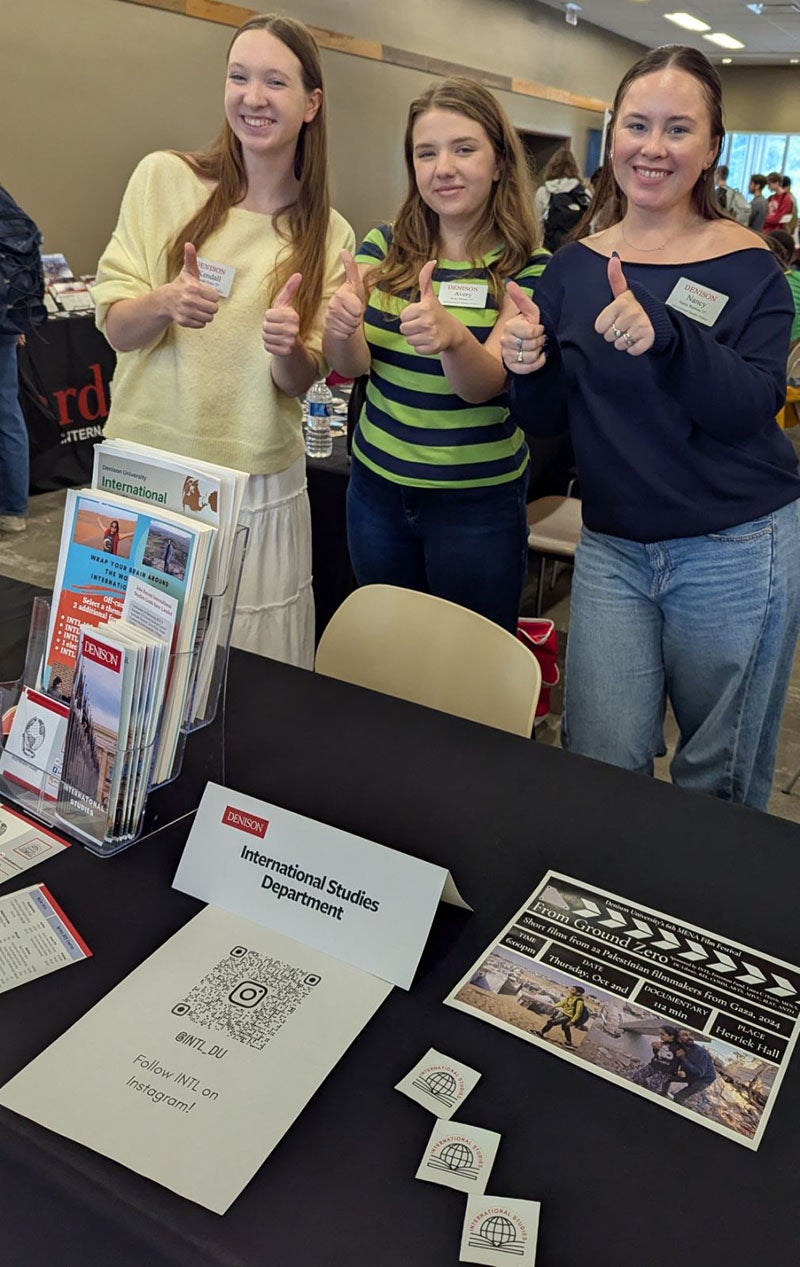 Three women behind an information table smile and offer a "thumbs up" to the camera.