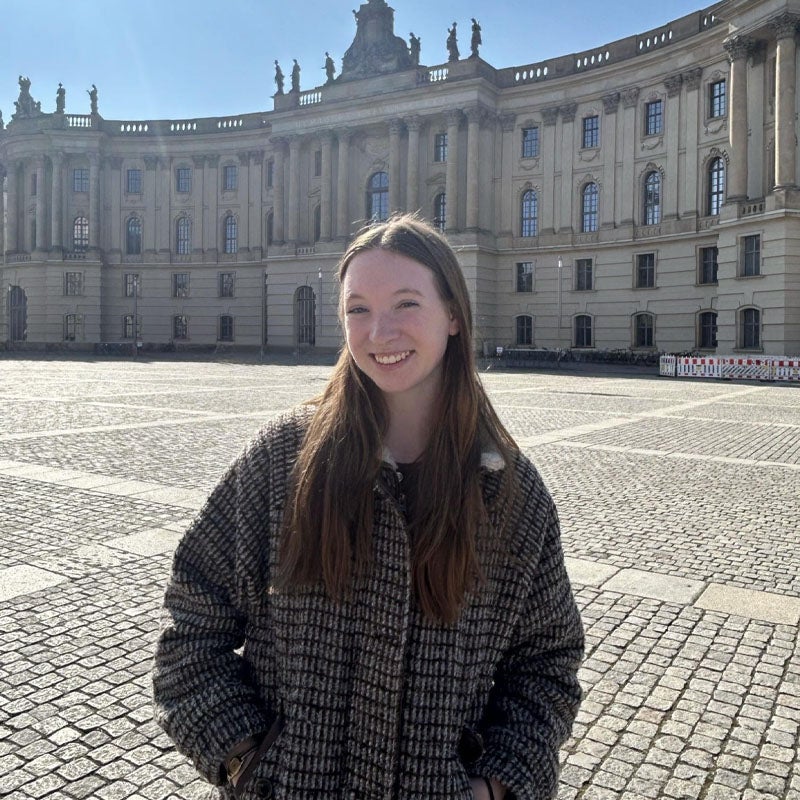 Kendall Polen A young woman with long brown hair stands smiling in a large, sunlit cobblestone plaza in front of a historic, curved building.