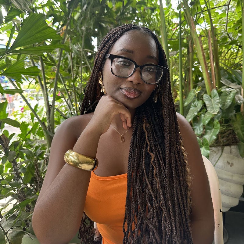 Kendrah Mpare A woman with long braids and glasses, wearing an orange top and gold jewelry, sits at a table in a lush, green outdoor setting.