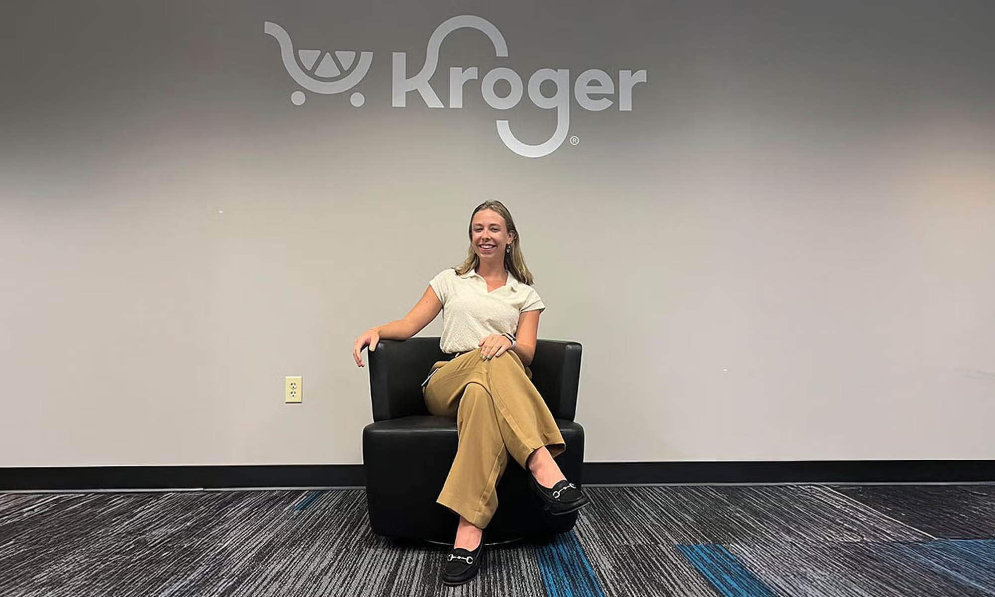Photo of a blonde-haired woman in a cream blouse and khaki pants sitting in a corporate office beneath a Kroger logo.