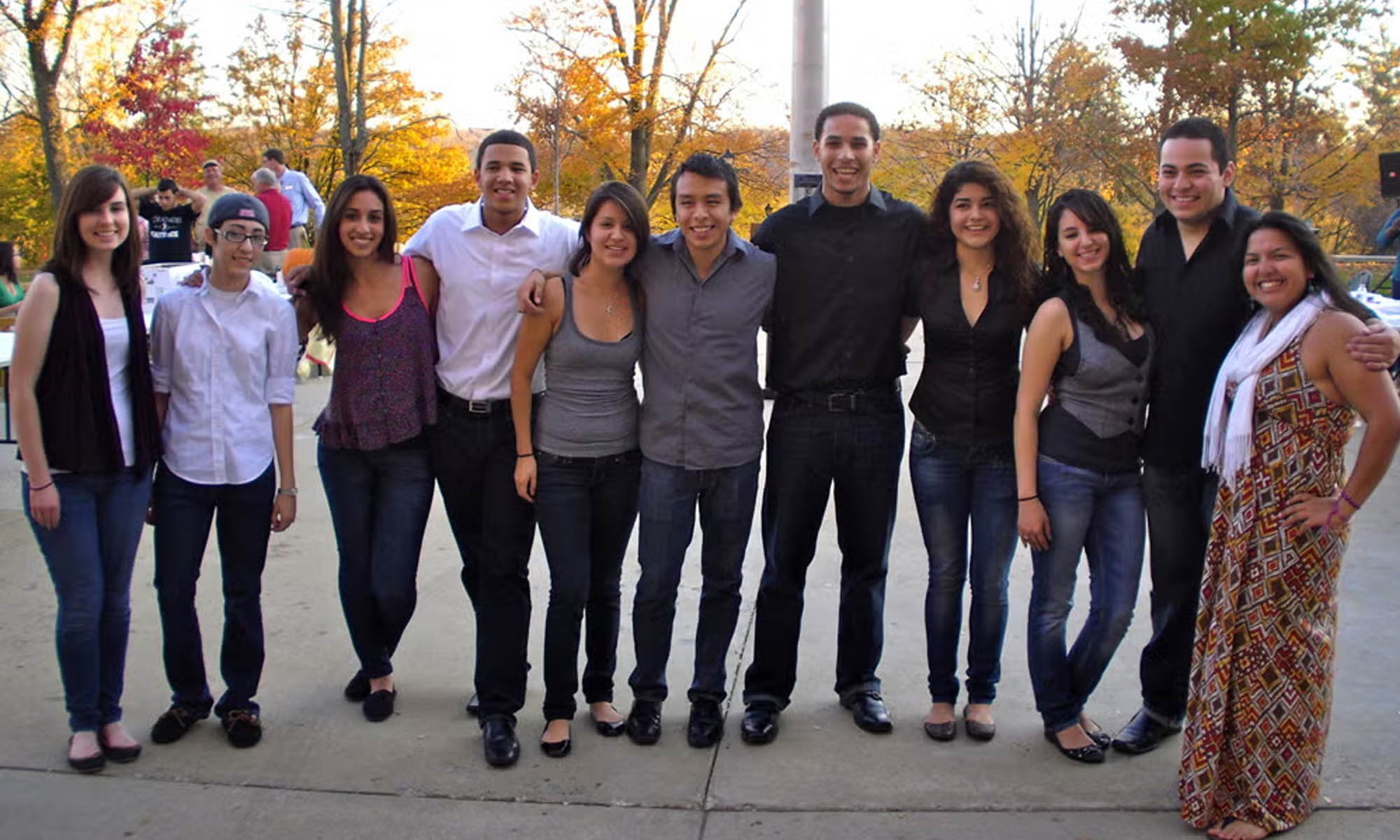 A group of eleven students stand in a line outside on a fall day, all smiling for the camera.