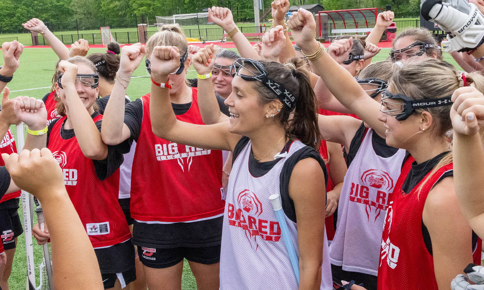 Women's lacrosse players in Denison gear cheering with their fists in the air on the field.