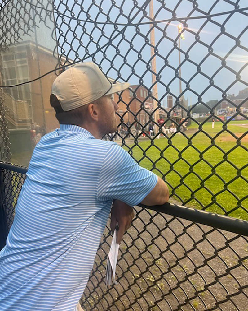 Photo of Matthew Zmuda looking at a baseball field through a fence along the sidelines.