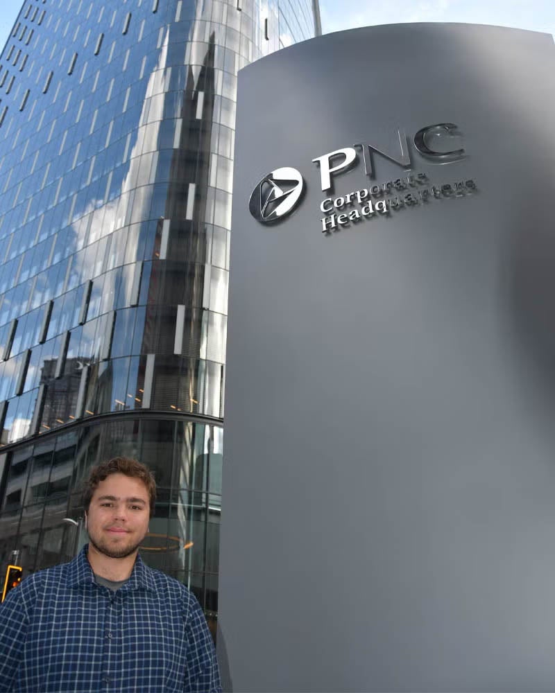 A young man with brown hair and a beard dressed in business casual clothes stands in front of tall, modern, silver and glass skyscraper next to a tall gray sign that says "PNC Corporate Headquarters"