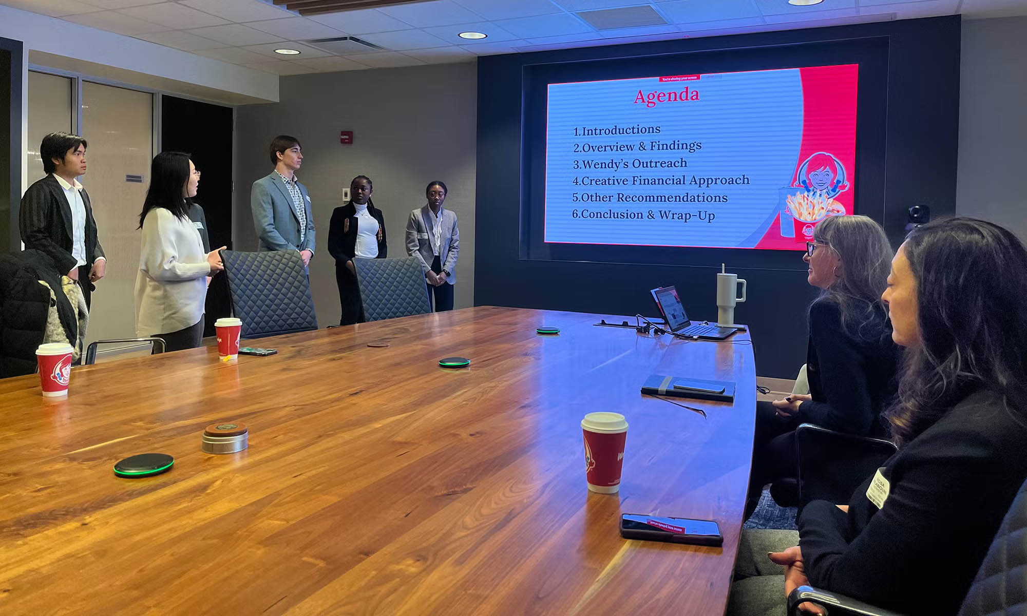 Five students in business wear making a presentation to executives in a conference room.