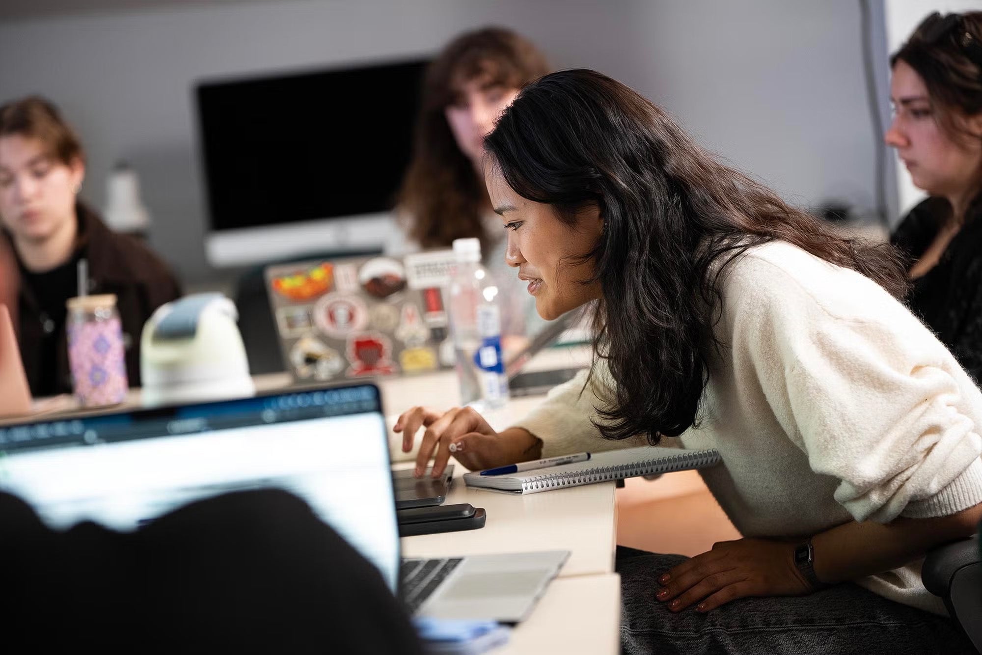 A woman sits at a desk using a laptop, surrounded by other people in an office setting.