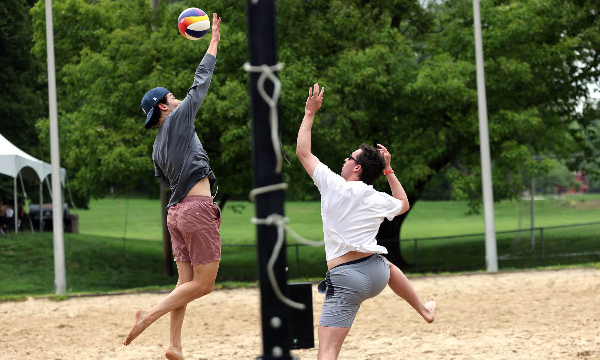Two students in shorts and tee shirts playing sand volleyball, one on each side of the net, both leaping up trying to get the volleyball between them.