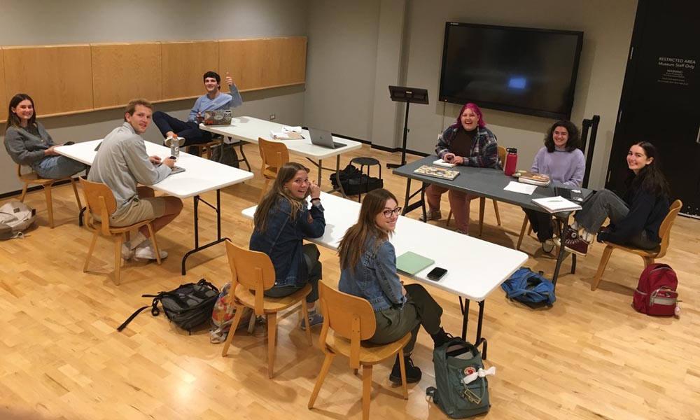 Wide angle photo of students sitting at tables in a meeting space, working on a project together.