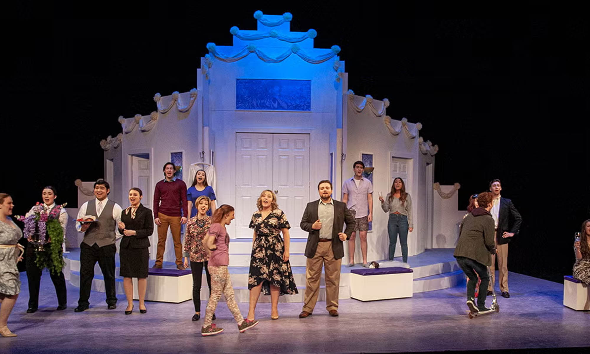 A cast of actors on a large stage in contemporary costumes standing in front of a large white set piece resembling a church shaped like a tiered wedding cake.