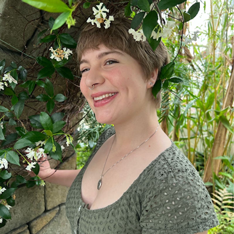 Sophia Scime A smiling woman in a green shirt stands near a wall that's covered in leaves, vines, and white flowers.